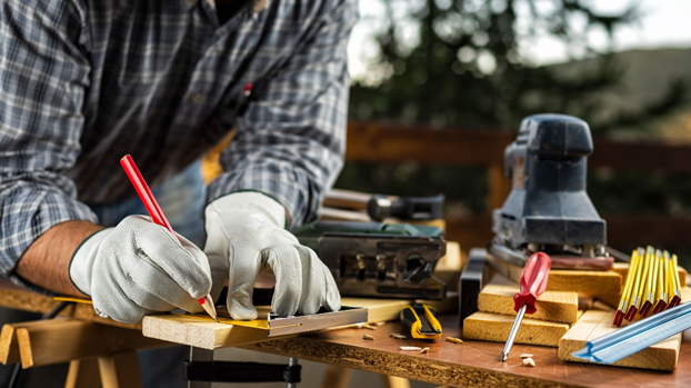 A person wearing gloves measures and marks a piece of wood with a pencil on a workbench, surrounded by various tools and pencils.