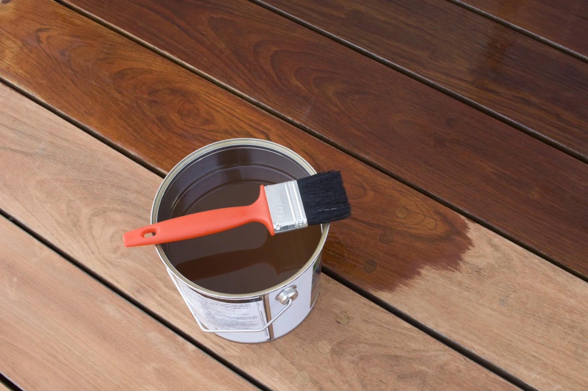 A paintbrush with a red handle rests on a can of wood stain on a wooden surface, with part of the wood already stained in a darker shade.