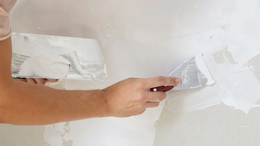 Close-up of a person using a trowel to spread plaster on a wall, smoothing the surface with a flat tool.