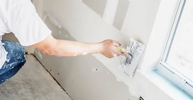 A person wearing a white shirt is using a trowel to apply plaster on a wall near a window.