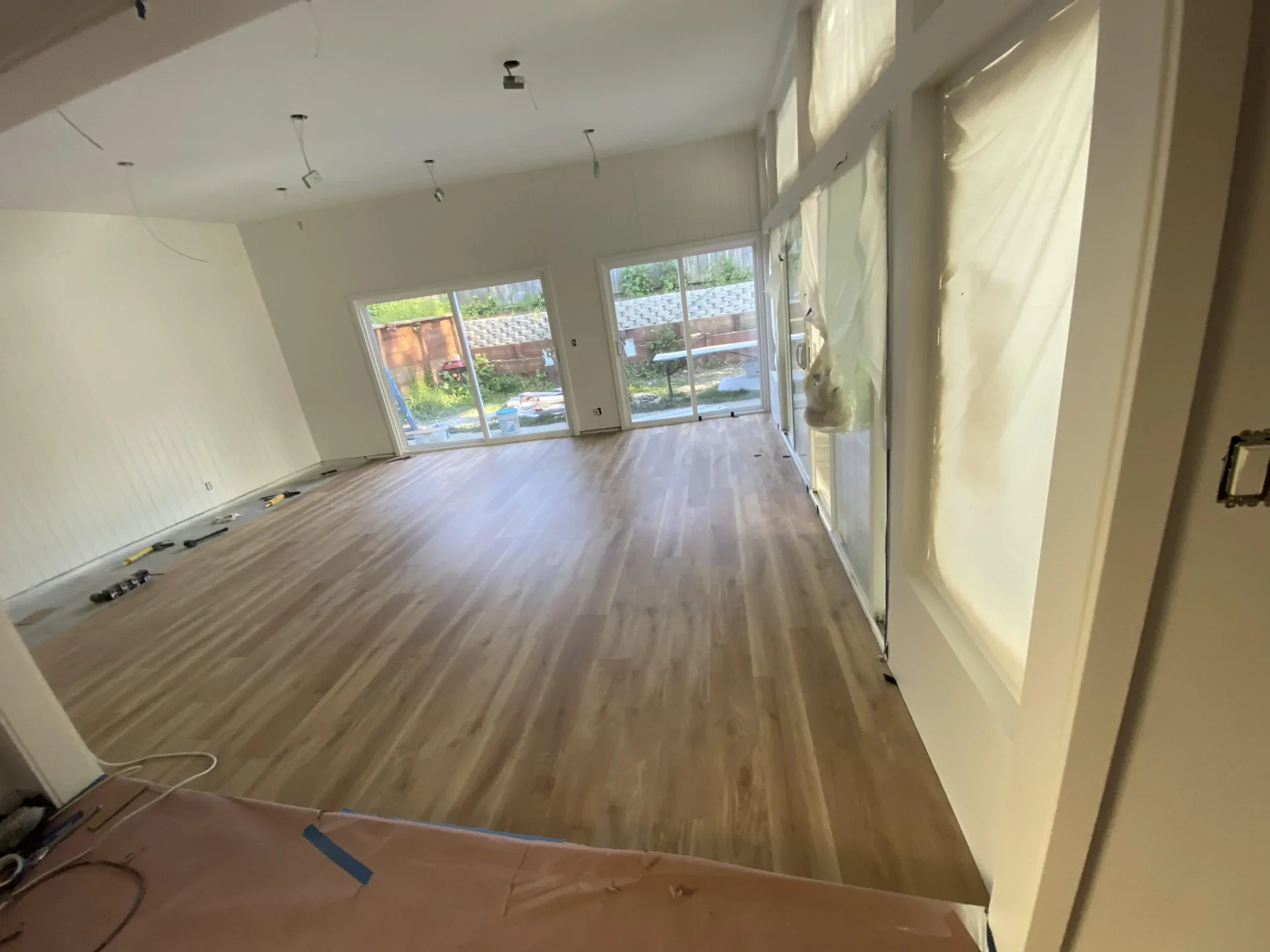 A room under renovation with newly installed light brown wood flooring, large windows, some covered with plastic, and partially unfinished walls and ceiling.