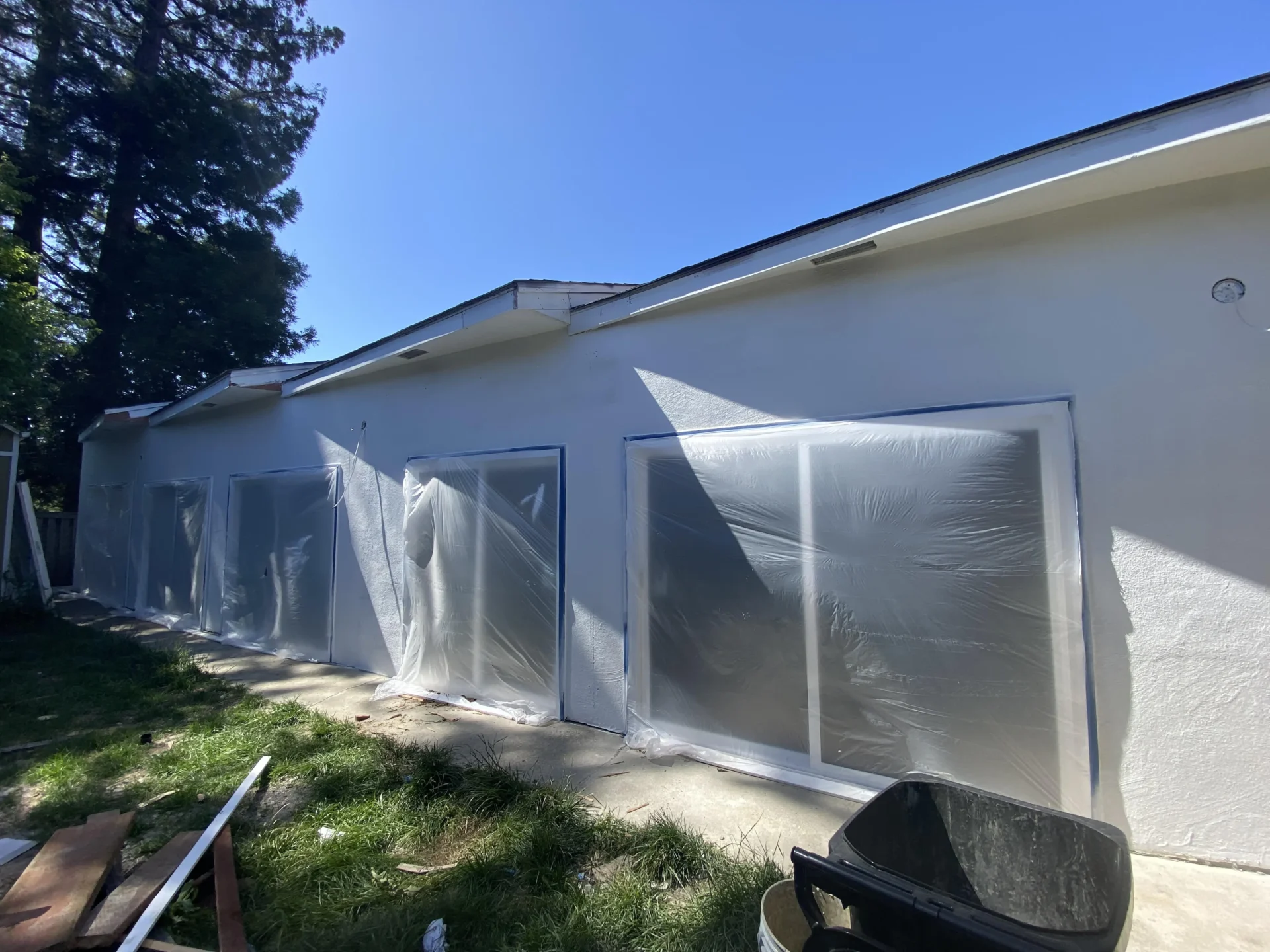 All-Pro-Painting-Projects-5 - All-Pro Painting Services A row of windows on a single-story building covered with plastic sheeting, part of an outdoor renovation project. A black wheelbarrow and some construction materials are in the foreground. exterior painting
