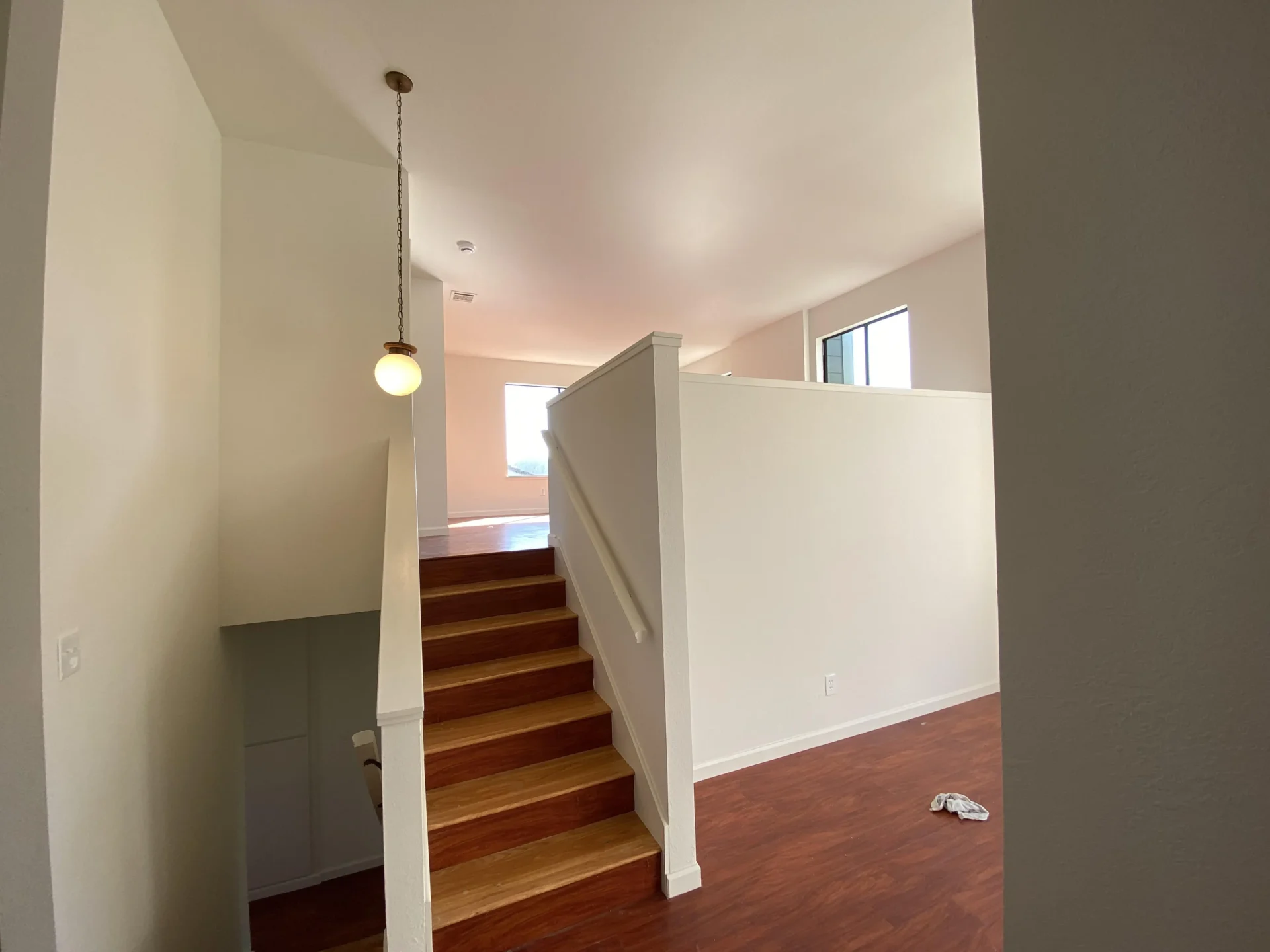 A well-lit interior showing wooden stairs leading up to a landing area with a white railing and a round light fixture hanging from the ceiling.