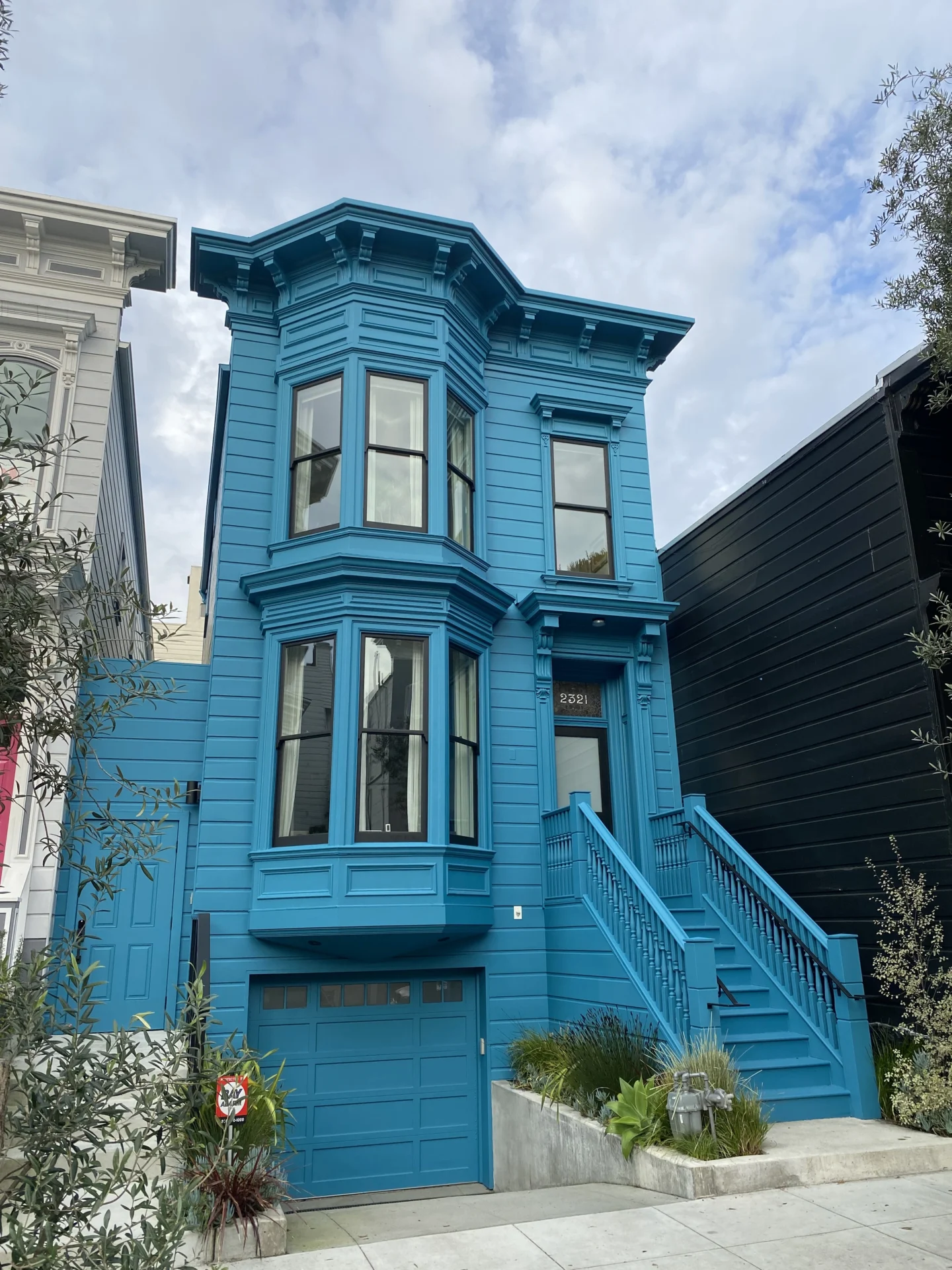 A two-story blue Victorian-style house with bay windows and a garage door underneath a staircase leading to the front door, set against a partly cloudy sky.