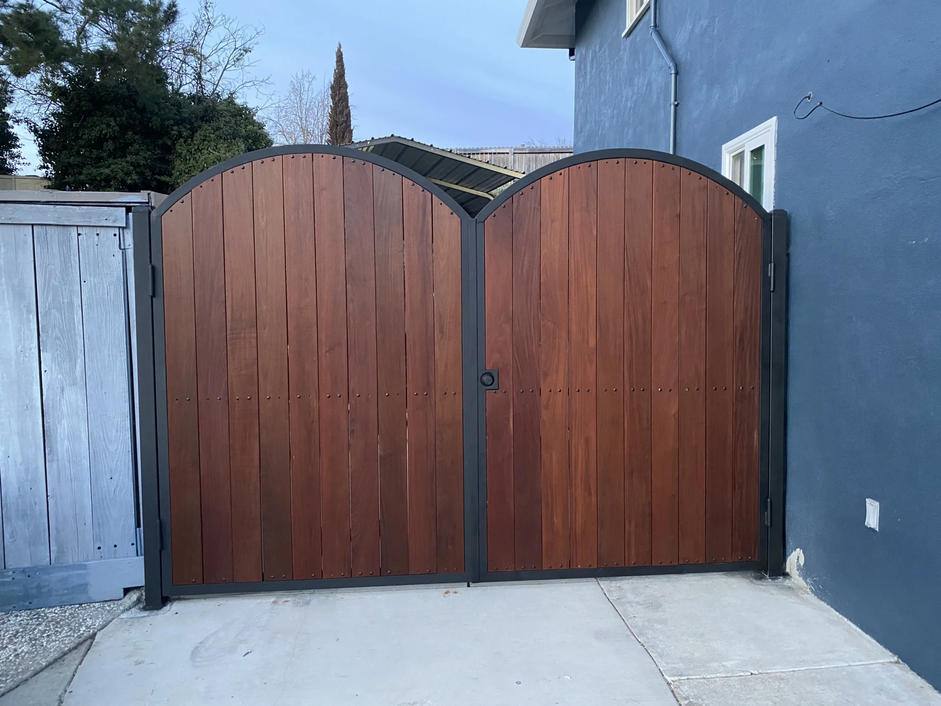 A wooden double gate with a curved top sits closed in front of a blue house, supported by black metal frames on both sides. A concrete path leads up to the gate.