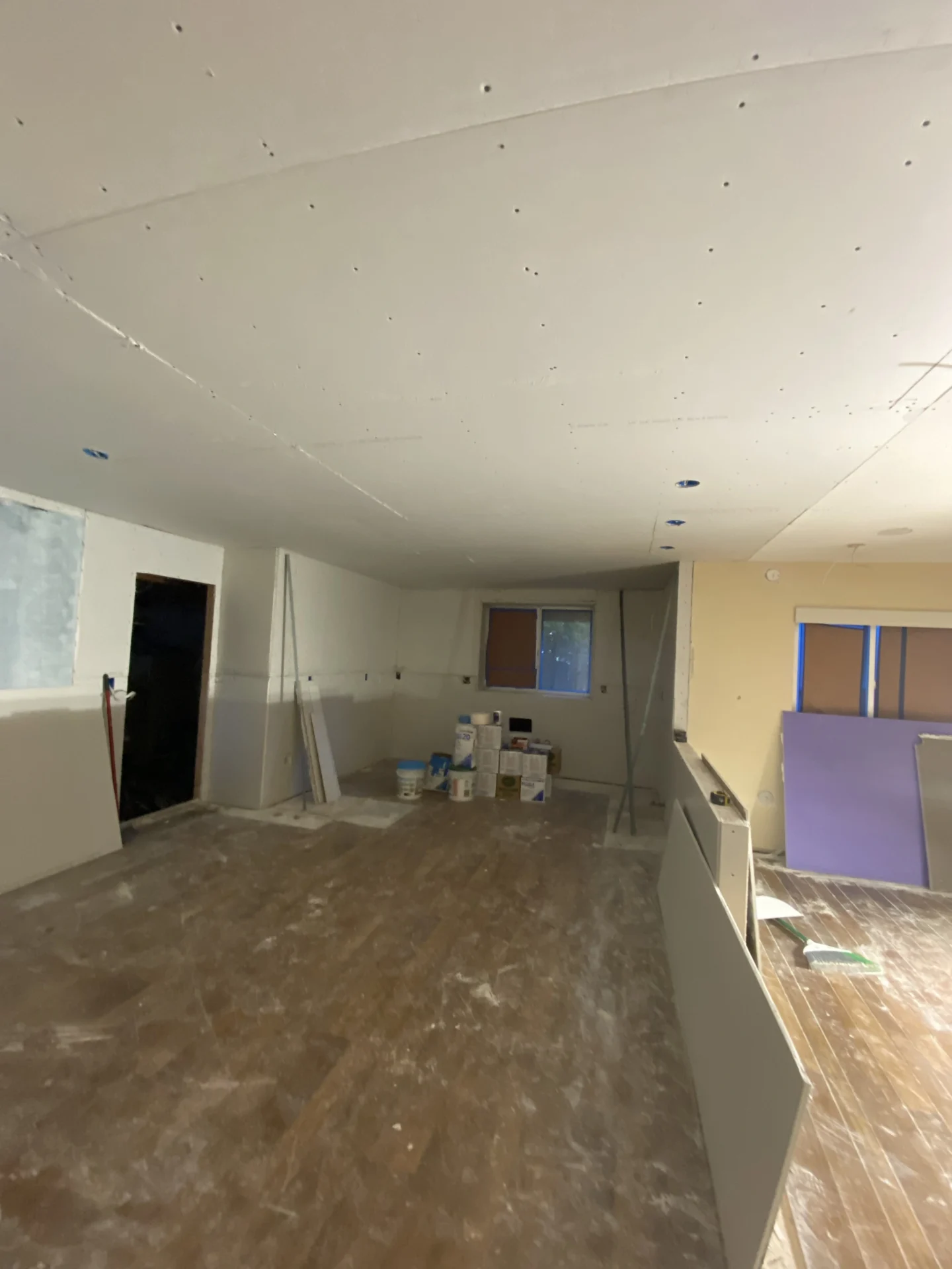A room under construction with drywall partially installed on walls and ceiling. The floor is covered in building materials, and windows are partially covered. Light fixtures are yet to be installed.