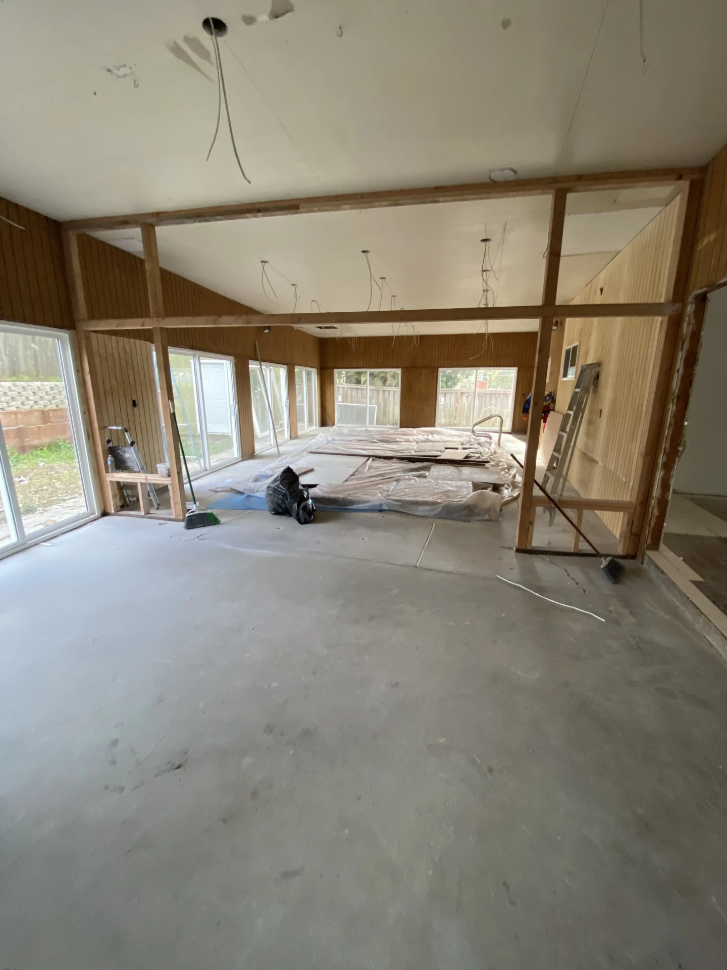 A large room under renovation, featuring exposed wooden framing, tools on the floor, and covered furniture. Walls with wooden panels and multiple windows provide natural light.