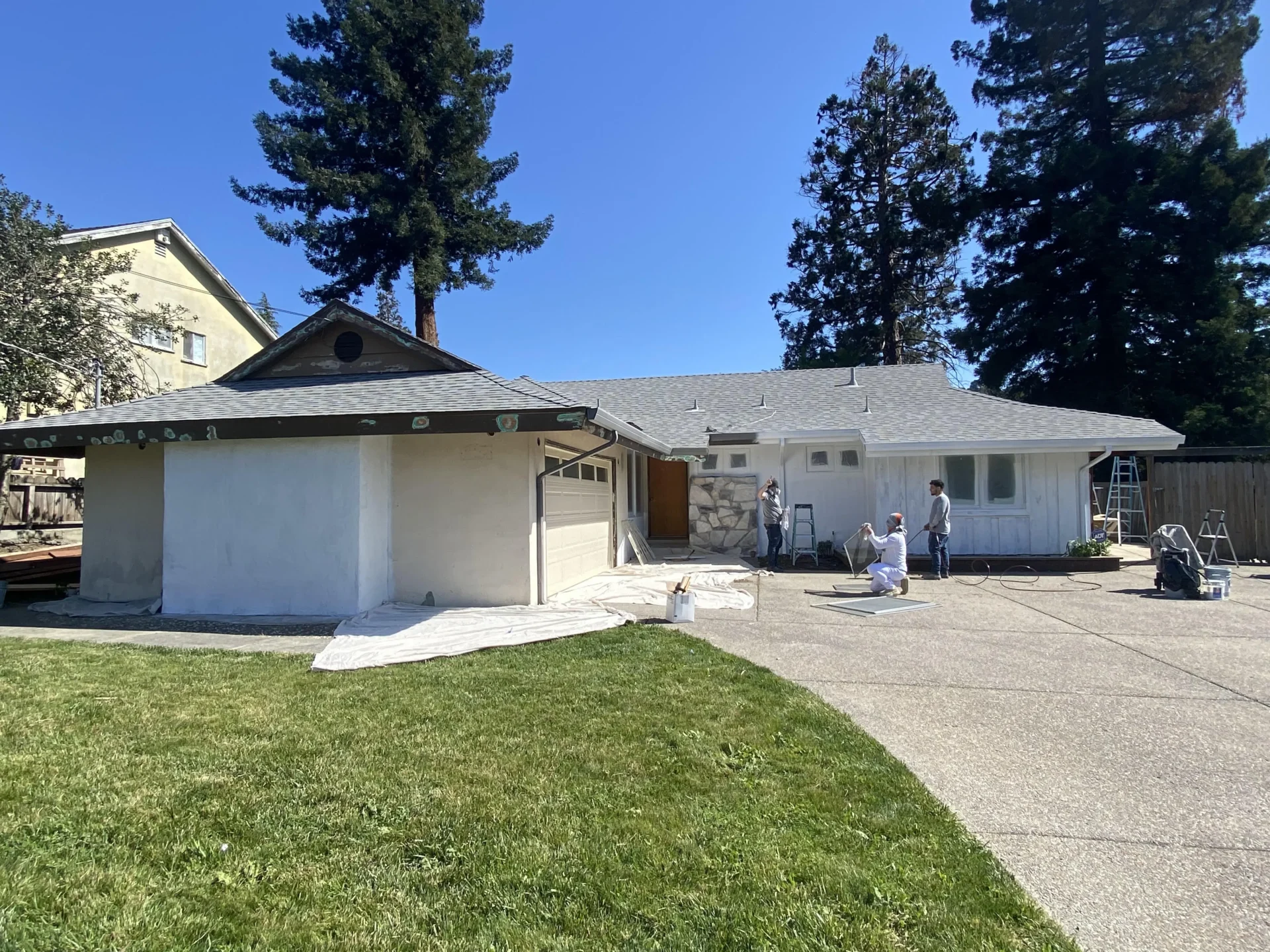 Workers painting the exterior of a single-story house under clear skies, with ladders and buckets visible on a paved driveway. A lawn is in the foreground.