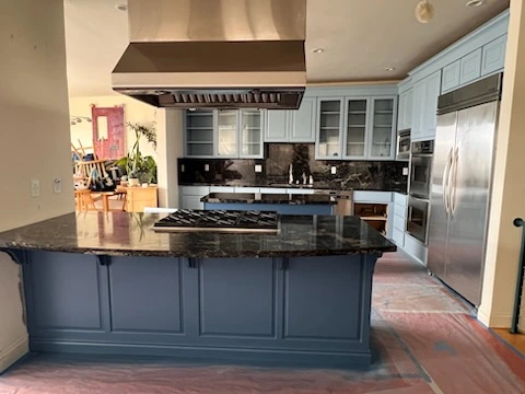 A modern kitchen features blue cabinets, a large island with a built-in stove, and stainless steel appliances, including a double-door refrigerator. The floor is covered with protective material.