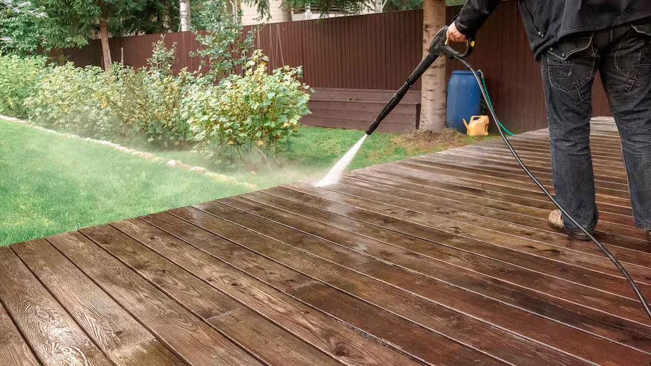 All-Pro-Power-Washing-2 - All-Pro Painting Services Person power washing a wooden deck in a backyard with green plants and a brown fence in the background.