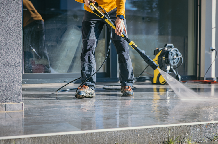 Person in black pants and orange jacket using a pressure washer to clean a tiled outdoor surface. A pressure washer machine is visible in the background.