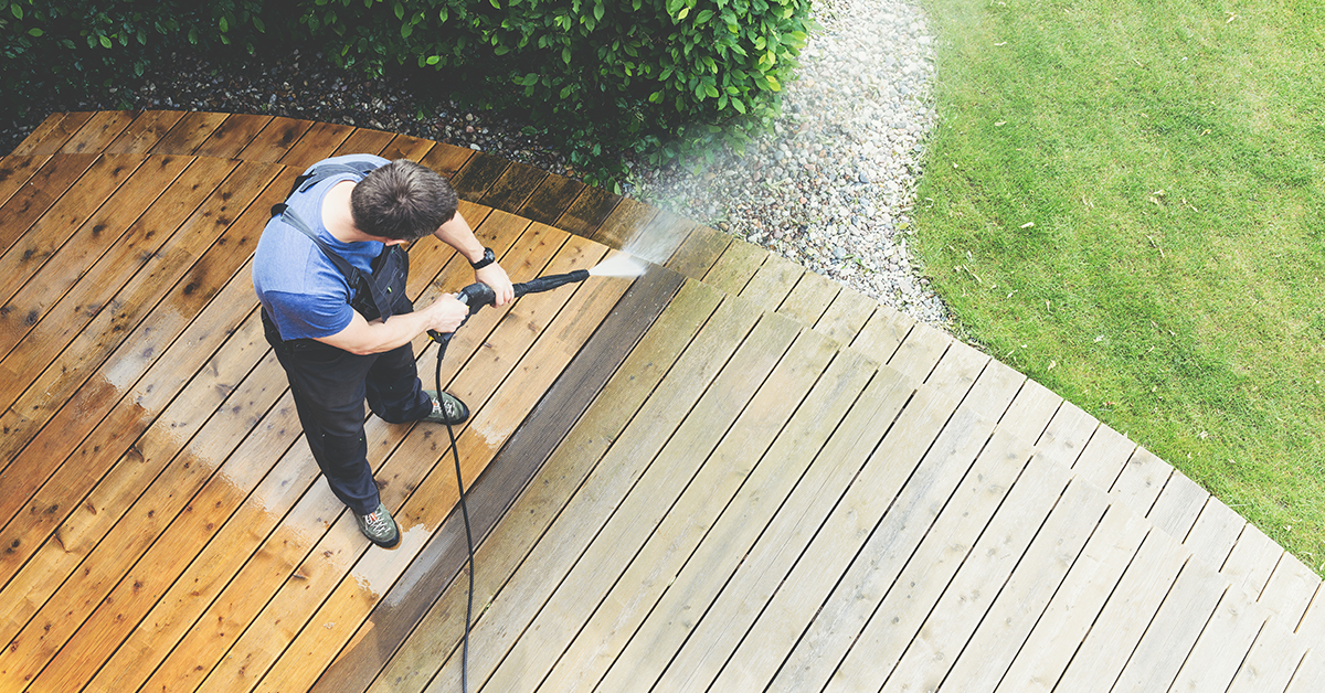 Person in overalls using a pressure washer to clean a wooden deck, with water visibly spraying and the difference between cleaned and uncleaned areas noticeable.