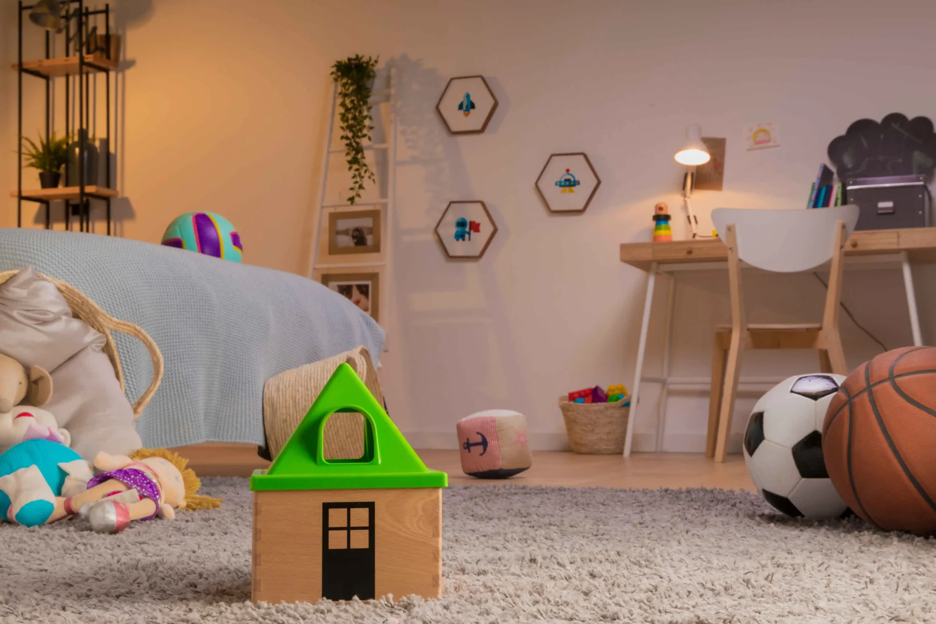A child’s bedroom features a bed, toys, a study desk with a lamp, and shelves with hexagonal wall decor. A basketball and soccer ball are on the floor next to a toy house.