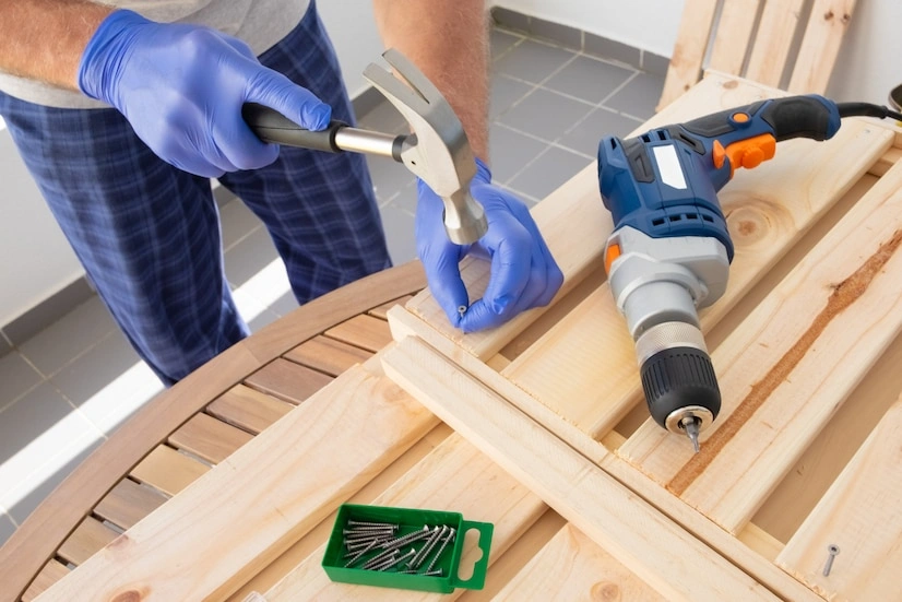 A person wearing blue gloves is using a hammer to drive a nail into a wooden board on a table. A cordless drill and a box of nails are also on the table.