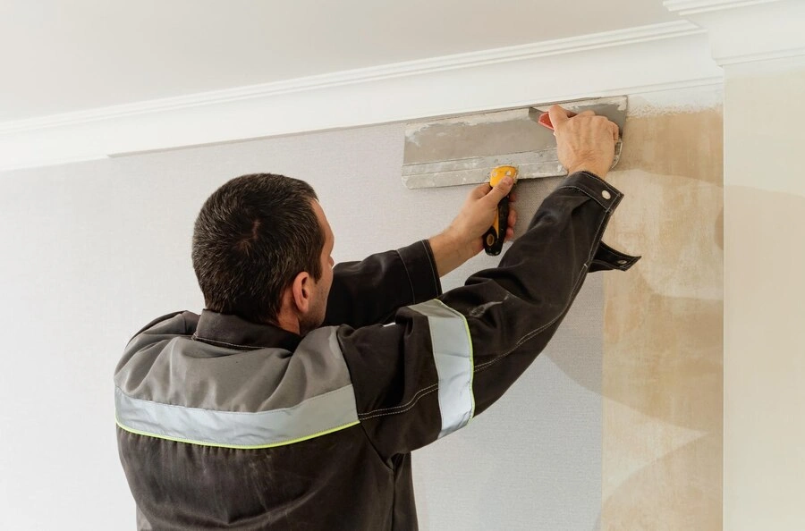 A person wearing a black and grey jacket uses a putty knife to apply plaster to a wall near the ceiling.