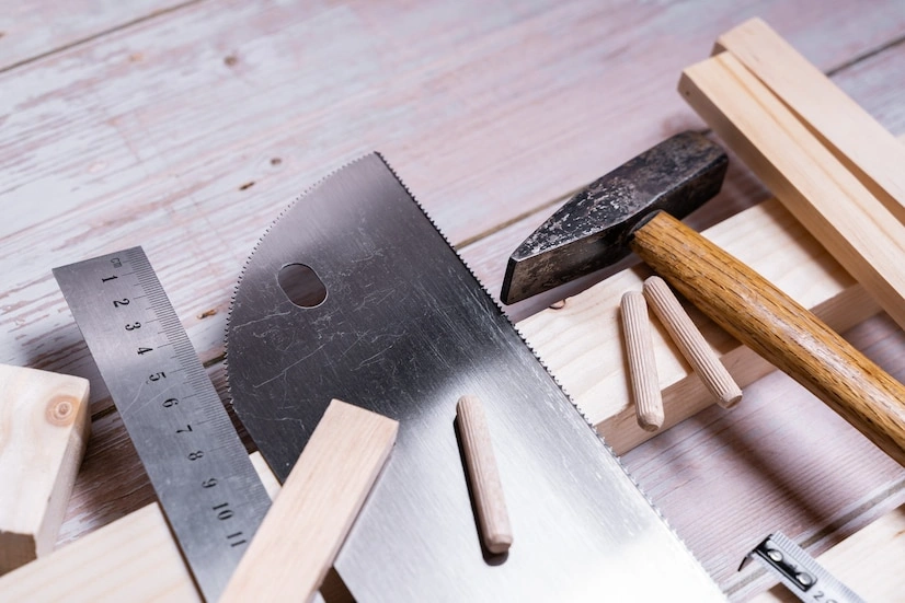 Woodworking tools including a saw, hammer, and ruler are arranged on a wooden surface along with several wooden dowels and planks.