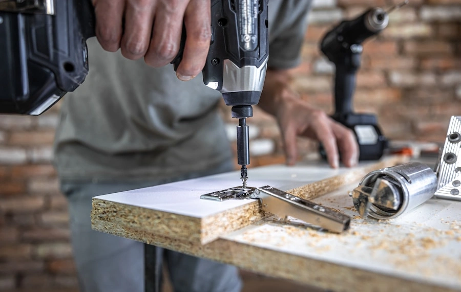 Person using a power drill to attach a hinge to a wooden board, with another power tool and metal parts on the workbench.