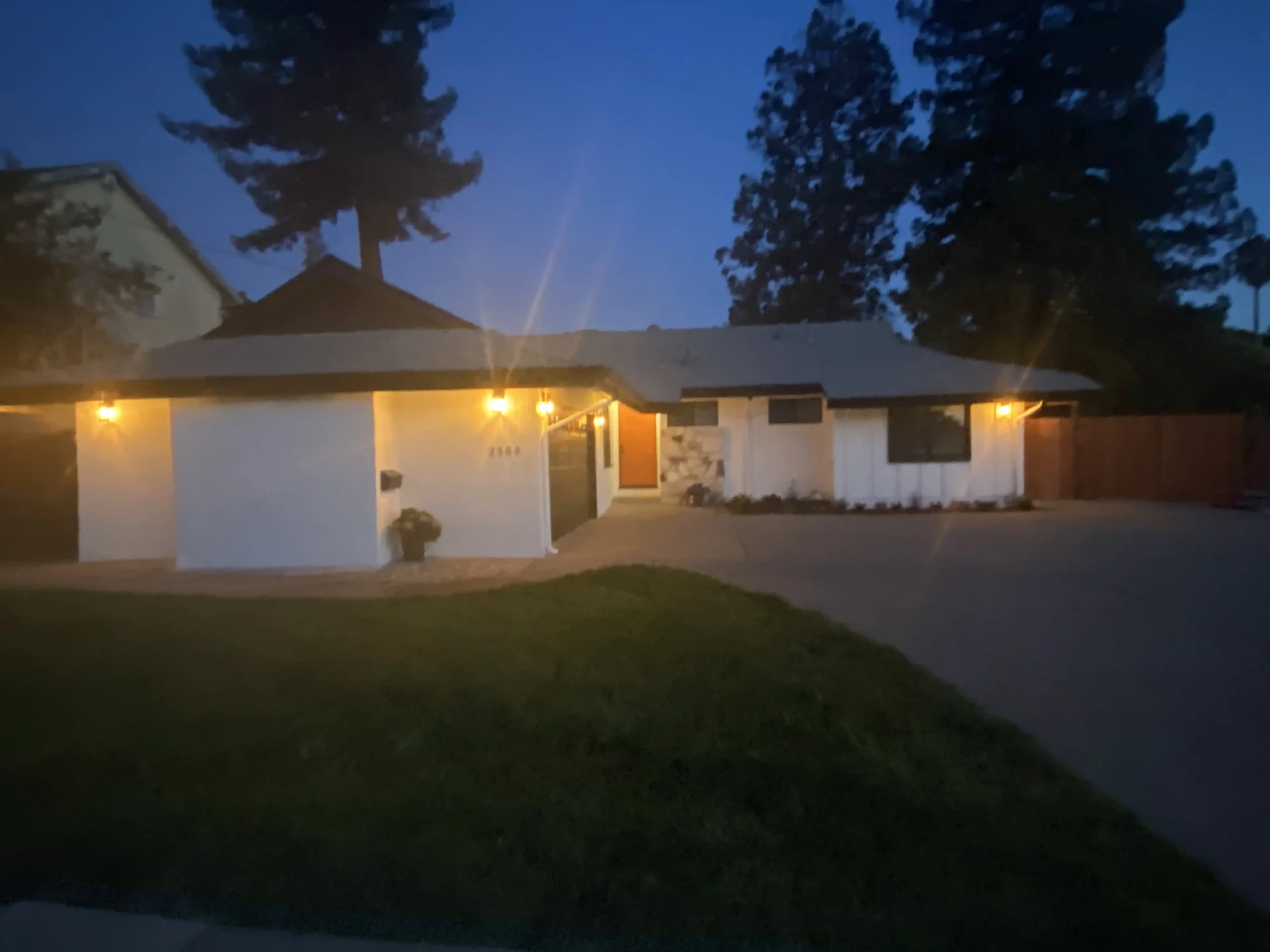 A single-story house at dusk with exterior lights on, surrounded by a lawn and trees. The house has a white facade, a peaked roof, and a concrete driveway.