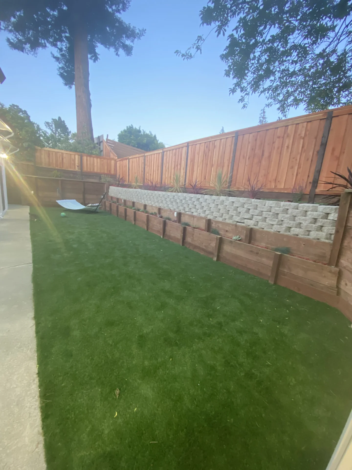 Backyard with artificial turf, concrete path on the left, and tiered wooden and stone retaining walls with plants, surrounded by a wooden fence.