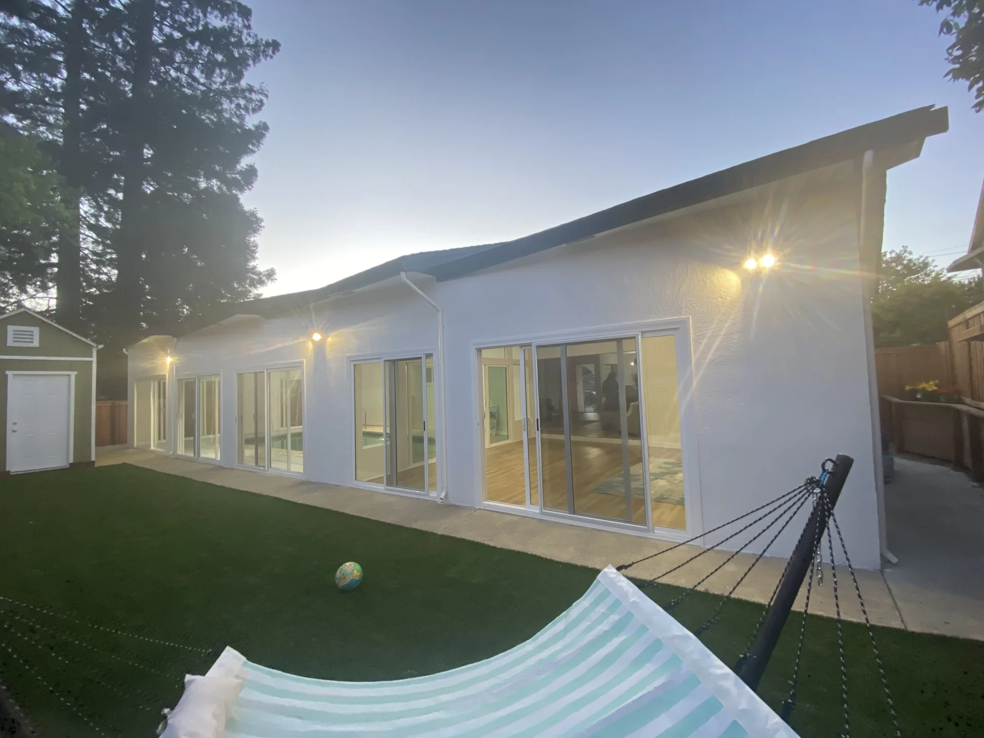 All-Pro-Painting-Exterior-Painting-9 - All-Pro Painting Services A modern white house with large sliding glass doors and outdoor lights illuminated in the evening. There's a hammock in the foreground on a grassy area near a small shed.