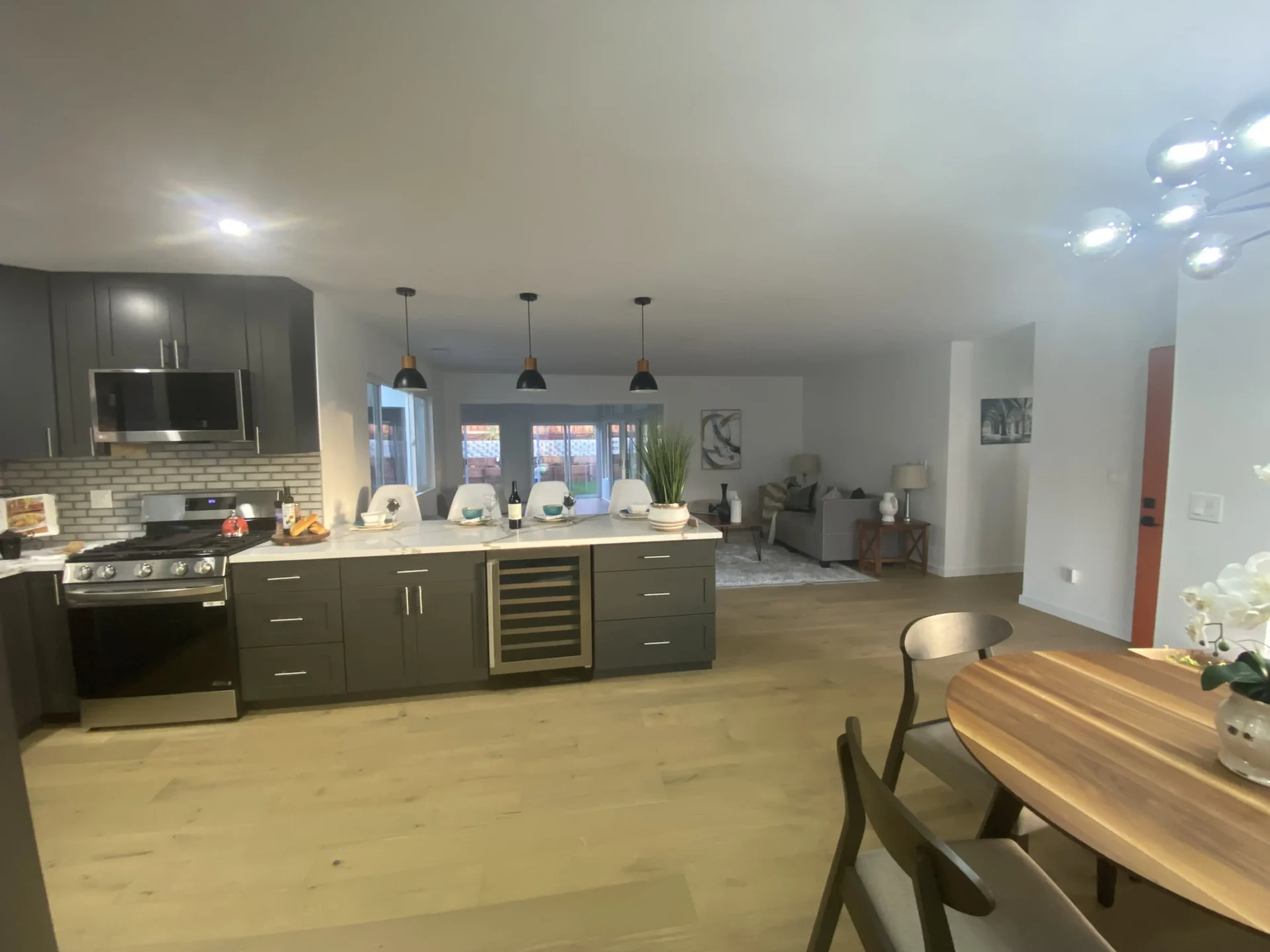 Modern open-concept kitchen and living area with dark cabinetry, a kitchen island with bar stools, a dining table, and a well-lit living room in the background.